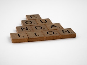Wooden Scrabble tiles are arranged to spell the word 'FOUNDATION' in a pyramid shape on a plain white background.