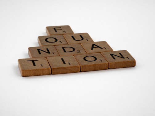 Wooden Scrabble tiles are arranged to spell the word 'FOUNDATION' in a pyramid shape on a plain white background.