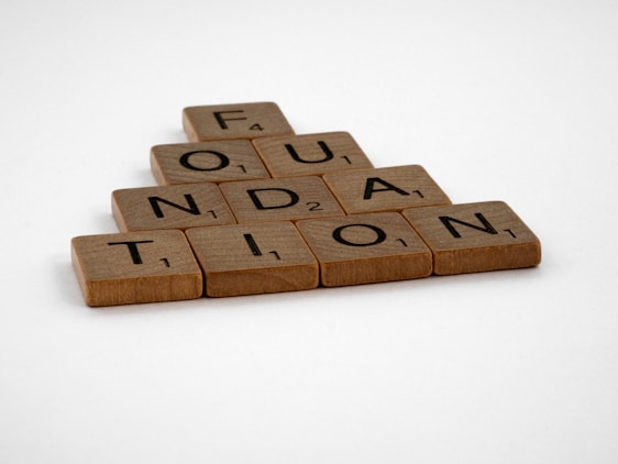 Wooden Scrabble tiles are arranged to spell the word 'FOUNDATION' in a pyramid shape on a plain white background.