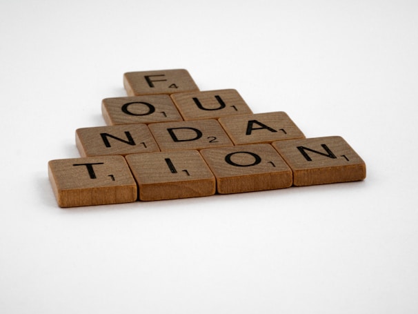 Wooden Scrabble tiles are arranged to spell the word 'FOUNDATION' in a pyramid shape on a plain white background.