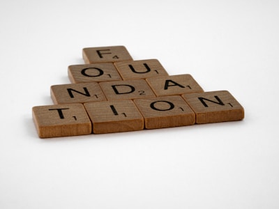 Wooden Scrabble tiles are arranged to spell the word 'FOUNDATION' in a pyramid shape on a plain white background.