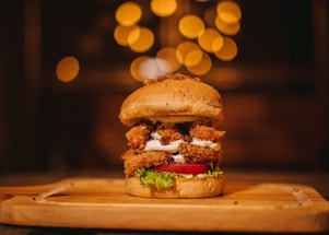 A gourmet burger is placed on a wooden serving board. It features a sesame seed bun filled with crispy, breaded chicken, fresh lettuce, a slice of ripe tomato, and a layer of white sauce. The background is softly blurred with warm, golden bokeh lighting, creating an inviting and cozy atmosphere.