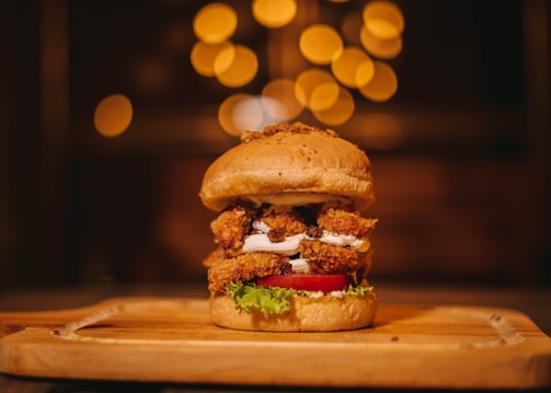 A gourmet burger is placed on a wooden serving board. It features a sesame seed bun filled with crispy, breaded chicken, fresh lettuce, a slice of ripe tomato, and a layer of white sauce. The background is softly blurred with warm, golden bokeh lighting, creating an inviting and cozy atmosphere.