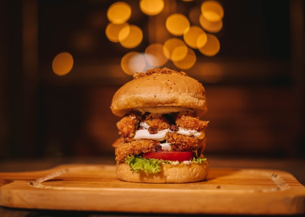 A gourmet burger is placed on a wooden serving board. It features a sesame seed bun filled with crispy, breaded chicken, fresh lettuce, a slice of ripe tomato, and a layer of white sauce. The background is softly blurred with warm, golden bokeh lighting, creating an inviting and cozy atmosphere.