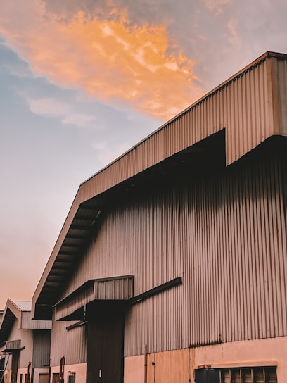 A large industrial building with a metal corrugated structure is pictured against a dramatic sky. The building features multiple sections with angular roofs, and the upper portion is bathed in warm, orange light from the setting or rising sun. The sky shows a contrast between warm, colorful clouds and the cooler, pale blue sky.