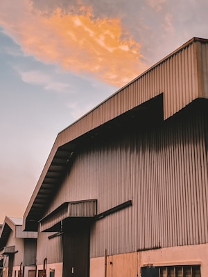 A large industrial building with a metal corrugated structure is pictured against a dramatic sky. The building features multiple sections with angular roofs, and the upper portion is bathed in warm, orange light from the setting or rising sun. The sky shows a contrast between warm, colorful clouds and the cooler, pale blue sky.