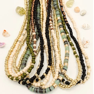 Multiple strands of colorful beads are arranged neatly, featuring a variety of bead types including gold, black, green, and cream hues. Small, decorative shells and stones are scattered around the beads on a light background.