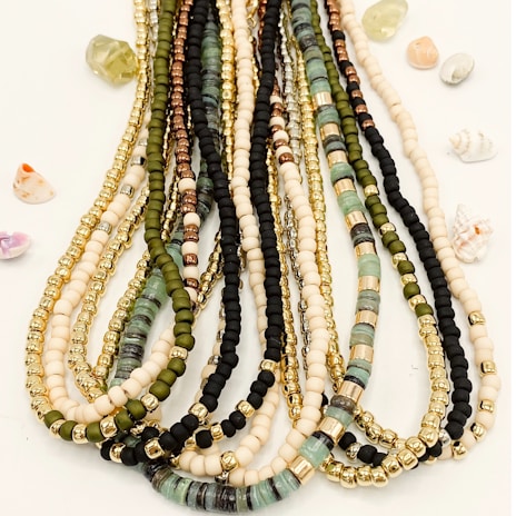 Multiple strands of colorful beads are arranged neatly, featuring a variety of bead types including gold, black, green, and cream hues. Small, decorative shells and stones are scattered around the beads on a light background.
