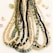 Multiple strands of colorful beads are arranged neatly, featuring a variety of bead types including gold, black, green, and cream hues. Small, decorative shells and stones are scattered around the beads on a light background.