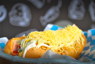 A loaded hot dog covered with shredded cheese, drizzled with mayonnaise and green sauce, is placed on a blue and white checkered paper lining a basket. The background features blurred illustrations of fish, suggesting a restaurant or seafood-themed setting.