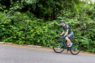 A cyclist wearing a helmet and sports attire is riding a road bike along a paved path bordered by lush green foliage. The setting conveys a sense of movement and outdoor activity, with the cyclist positioned against a backdrop of dense forest vegetation.