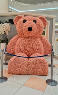A large teddy bear made entirely of pink roses is displayed in a public indoor setting. The bear is roped off with a blue cord and is surrounded by polished tile flooring. Nearby, there are some white orchids, and the environment appears to be well-lit with modern lighting.