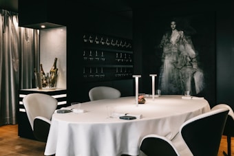 A sophisticated dining room featuring a round table with a white tablecloth, modern white and black chairs, and minimalistic table settings. On the table, a small floral centerpiece and two sleek, modern lamps provide a touch of elegance. To the left, a metallic open shelf displays wine bottles, and several wine glasses are hung. The backdrop is a dark wall with a large, artistic portrait that adds a sense of mystery and style. Soft, shimmering curtains partially cover a window, enhancing the ambience.