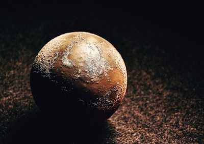 A spherical object with a textured surface that resembles a planet. The surface features craters and uneven patterns, illuminated by a warm light source against a dark background.