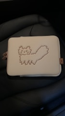 A cream-colored rectangular pouch featuring a stitched outline of a cat in a playful pose. The pouch lies on a dark cushioned surface, likely a car seat, and includes a small side tag.