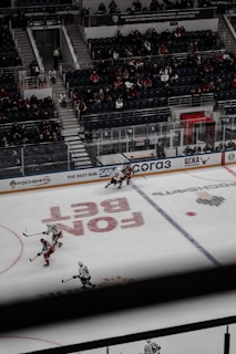 A hockey game taking place in an indoor arena with a partially filled audience watching from the stands. Players from two teams, wearing white and red jerseys, are engaged in the game on the ice rink. The arena features advertisements along the borders of the rink.