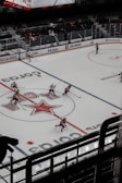A professional ice hockey game is underway in an arena with players actively skating on the ice. The rink features prominent logos and advertisements. Players are wearing uniforms in red and white, and an electronic scoreboard is visible above the stands. Sparse spectators are sitting behind glass barriers.