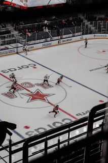 A professional ice hockey game is underway in an arena with players actively skating on the ice. The rink features prominent logos and advertisements. Players are wearing uniforms in red and white, and an electronic scoreboard is visible above the stands. Sparse spectators are sitting behind glass barriers.