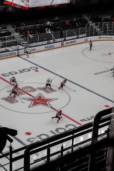 A professional ice hockey game is underway in an arena with players actively skating on the ice. The rink features prominent logos and advertisements. Players are wearing uniforms in red and white, and an electronic scoreboard is visible above the stands. Sparse spectators are sitting behind glass barriers.