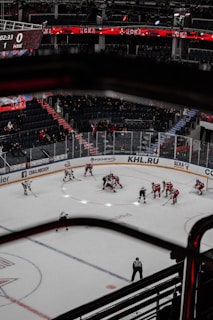 An ice hockey game is taking place in a large indoor arena. Players are positioned for a face-off at center ice, with teams wearing red and white uniforms. The arena features dark seating areas, some occupied by spectators. Overhead, a digital scoreboard displays the score and time. There are prominent advertisements along the boards, and bright lights illuminate the ice.