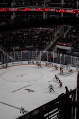 A hockey game is in progress in a large arena with players actively moving around the ice. The stands have a moderate number of spectators. Team players in red and white uniforms are engaged in the match near one of the goals, with some players attempting to control the puck. Overhead, digital screens display information in red and white.