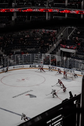 A hockey game is in progress in a large arena with players actively moving around the ice. The stands have a moderate number of spectators. Team players in red and white uniforms are engaged in the match near one of the goals, with some players attempting to control the puck. Overhead, digital screens display information in red and white.