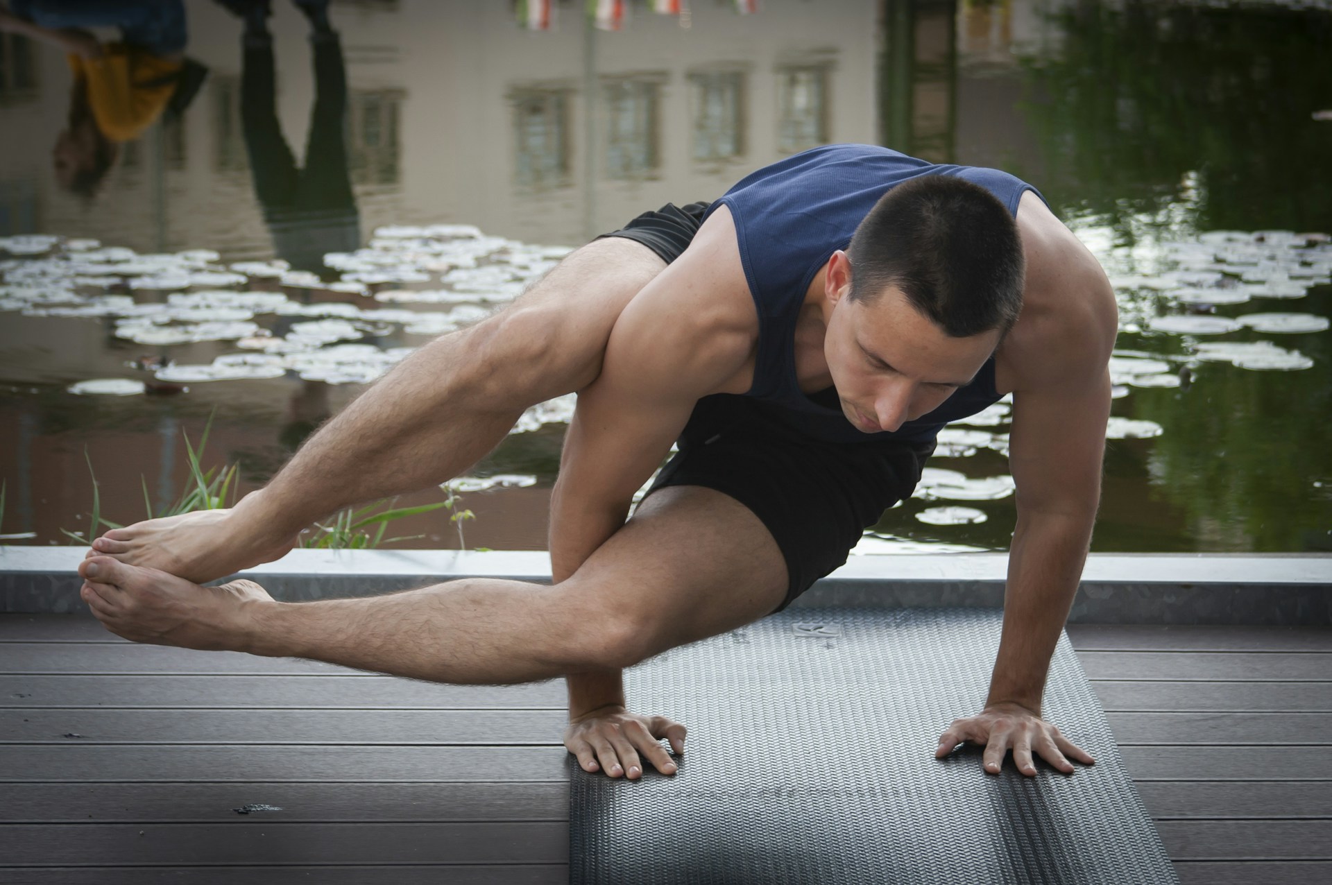 A man is practicing a yoga or acrobatic pose on a dark exercise mat near a pond with water lilies. His body is balanced on one hand while his legs are elevated and crossed, showing strength and concentration. The background reflects the surroundings in the water, with lush greenery visible.