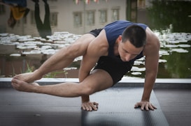 A man is practicing a yoga or acrobatic pose on a dark exercise mat near a pond with water lilies. His body is balanced on one hand while his legs are elevated and crossed, showing strength and concentration. The background reflects the surroundings in the water, with lush greenery visible.