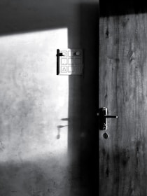 A wooden door is partially open, casting a shadow on the adjacent wall. Sunlight streams in, highlighting a modern light switch panel on the wall. A keychain hangs from the door handle, visible in the shadow.