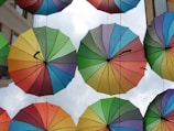 A vibrant display of colorful umbrellas suspended in the air, forming a canopy against a cloudy sky. Each umbrella features a spectrum of colors with a central black handle.