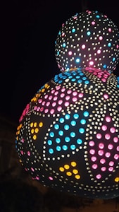 A spherical, intricately designed lamp with numerous small perforations emits vibrant colors. The patterns include multiple circular shapes filled with colorful lights including blue, pink, and yellow. Set against a dark background, the lamp creates an eye-catching display with its illuminated patterns.