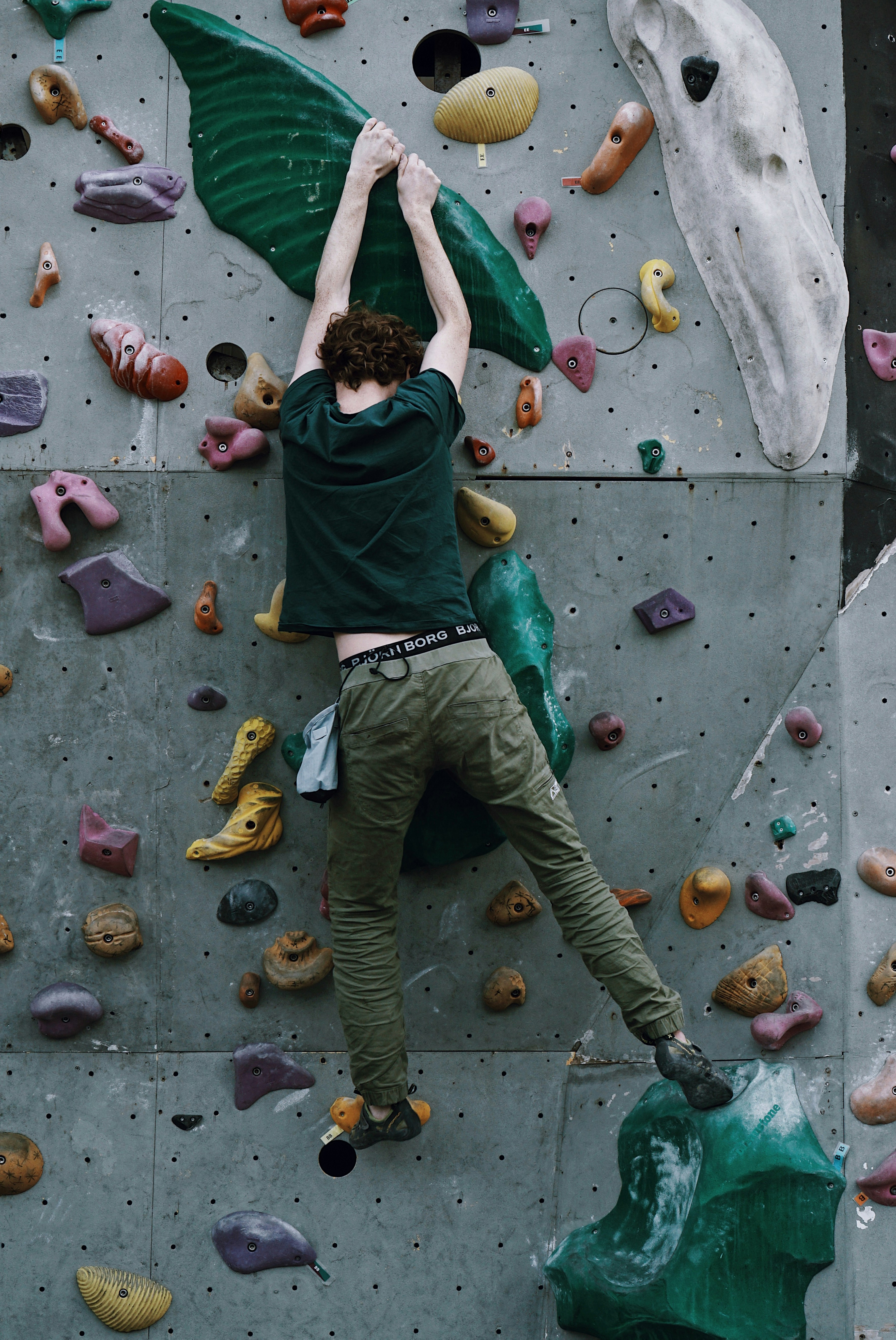 A person is climbing an indoor rock wall, utilizing various colorful grips and holds affixed to a textured vertical surface. The climber is wearing a dark green shirt and khaki pants, and a chalk bag is attached to their waist.