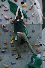 A person is climbing an indoor rock wall, utilizing various colorful grips and holds affixed to a textured vertical surface. The climber is wearing a dark green shirt and khaki pants, and a chalk bag is attached to their waist.