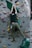 A person is climbing an indoor rock wall, utilizing various colorful grips and holds affixed to a textured vertical surface. The climber is wearing a dark green shirt and khaki pants, and a chalk bag is attached to their waist.