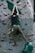A person is climbing an indoor rock wall, utilizing various colorful grips and holds affixed to a textured vertical surface. The climber is wearing a dark green shirt and khaki pants, and a chalk bag is attached to their waist.