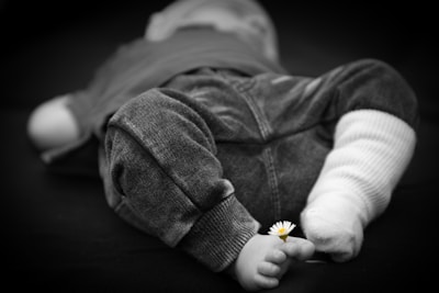 A black and white photograph of a baby lying down, focusing on the lower body. The baby is wearing soft pants and one sock, with a small daisy flower placed between the toes, providing a gentle contrast to the monochromatic tones.