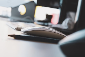 A modern, sleek computer mouse is placed on a desk, accompanied by a blurry glimpse of a keyboard in the background. The scene is minimally lit with natural light filtering in, giving it an airy feel. The focus is primarily on the mouse, with other elements such as a part of a monitor and peripheral items blurred out.