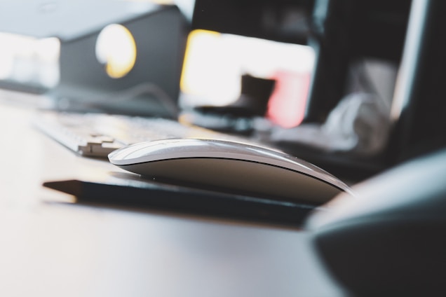 A modern, sleek computer mouse is placed on a desk, accompanied by a blurry glimpse of a keyboard in the background. The scene is minimally lit with natural light filtering in, giving it an airy feel. The focus is primarily on the mouse, with other elements such as a part of a monitor and peripheral items blurred out.
