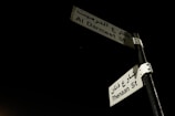 Two street signs are mounted on a metal pole against a dark night sky. The signs are labeled in both English and Arabic, displaying the names 'Al Darmeet St' and 'Thenaan St'. The signs are white with black text and are slightly illuminated, standing out against the dark background.