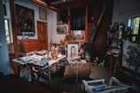 A cluttered artist's studio filled with various painting supplies, canvases, and artwork. An easel holds a portrait of a person, surrounded by paint tubes and brushes on a worktable. The room has a rustic feel with wooden beams, a brick wall, and a wood-burning stove. Books and other materials are stacked on shelves, contributing to the creative and slightly chaotic atmosphere.