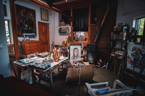 A cluttered artist's studio filled with various painting supplies, canvases, and artwork. An easel holds a portrait of a person, surrounded by paint tubes and brushes on a worktable. The room has a rustic feel with wooden beams, a brick wall, and a wood-burning stove. Books and other materials are stacked on shelves, contributing to the creative and slightly chaotic atmosphere.