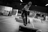 A person is performing a skateboard trick inside an indoor skate park. The area is filled with ramps and graffiti-covered walls. The setting has a raw, urban feel with its exposed architecture and dim lighting. The person's attire includes a hoodie and beanie, fitting the casual and gritty aesthetic typical of skate culture.