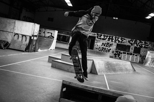 A person is performing a skateboard trick inside an indoor skate park. The area is filled with ramps and graffiti-covered walls. The setting has a raw, urban feel with its exposed architecture and dim lighting. The person's attire includes a hoodie and beanie, fitting the casual and gritty aesthetic typical of skate culture.