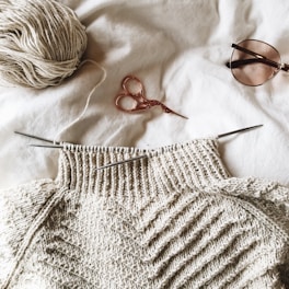 A partially knitted sweater made with cream-colored yarn is placed on a white surface. Next to it, there is a ball of yarn, a pair of small metallic scissors with an ornate handle, and a pair of sunglasses.