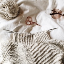 A partially knitted sweater made with cream-colored yarn is placed on a white surface. Next to it, there is a ball of yarn, a pair of small metallic scissors with an ornate handle, and a pair of sunglasses.