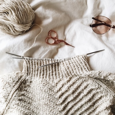 A partially knitted sweater made with cream-colored yarn is placed on a white surface. Next to it, there is a ball of yarn, a pair of small metallic scissors with an ornate handle, and a pair of sunglasses.