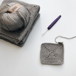 A crochet project in progress with a ball of gray yarn resting on a stack of two gray crochet pieces. A partially completed square crochet piece lies nearby. A purple crochet hook is positioned close to the yarn and crochet work.