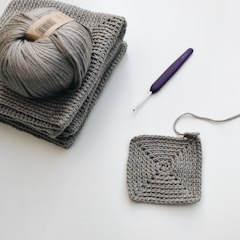 A crochet project in progress with a ball of gray yarn resting on a stack of two gray crochet pieces. A partially completed square crochet piece lies nearby. A purple crochet hook is positioned close to the yarn and crochet work.