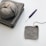 A crochet project in progress with a ball of gray yarn resting on a stack of two gray crochet pieces. A partially completed square crochet piece lies nearby. A purple crochet hook is positioned close to the yarn and crochet work.