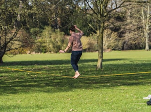 A person is balancing on a slackline in a park. They are barefoot and wearing casual clothes. Surrounding them are trees without leaves, and the ground is covered with green grass under a clear sky.